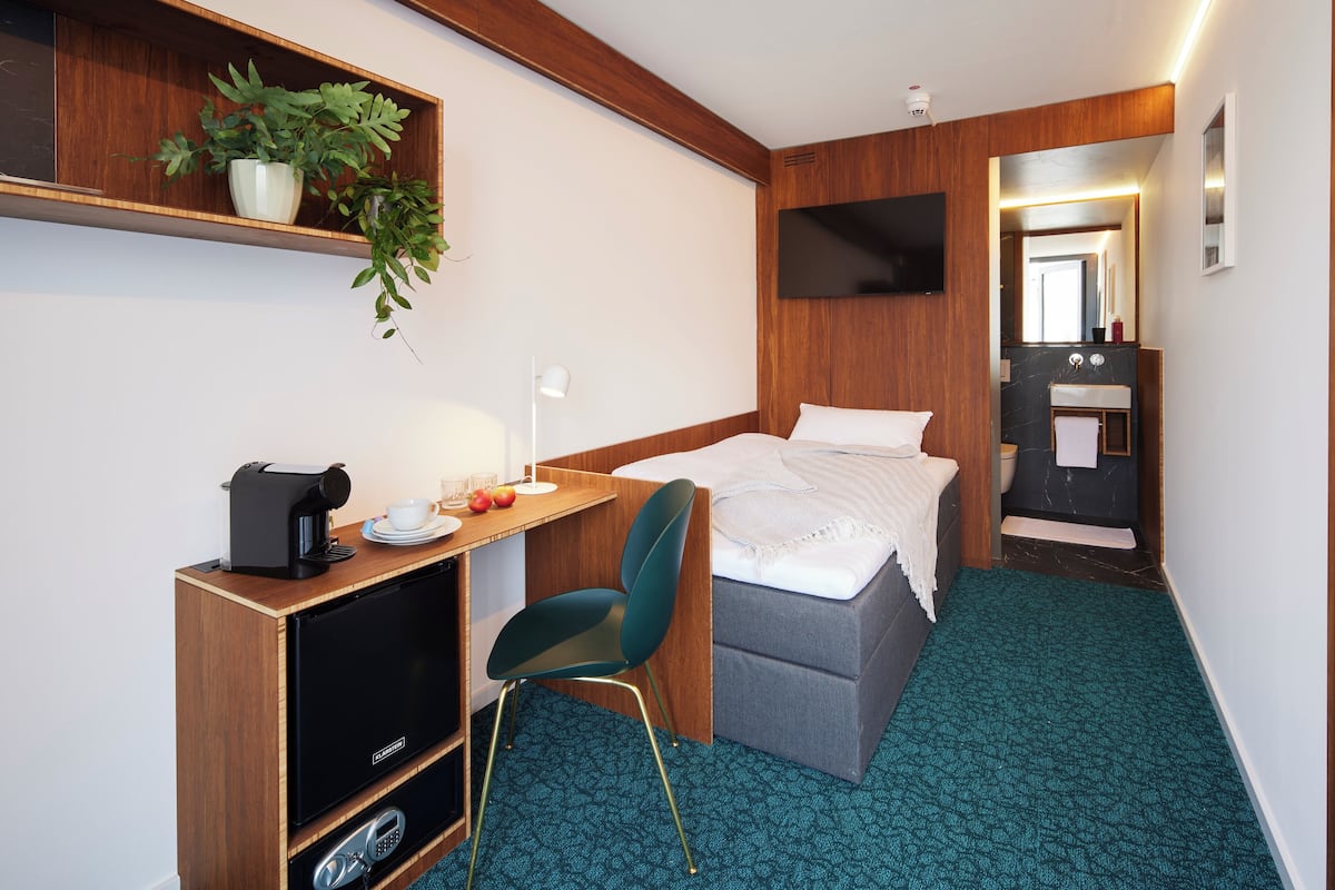 Una habitación de hotel compacta con cama, escritorio con cafetera, silla, televisor y baño con espejo y lavamanos.