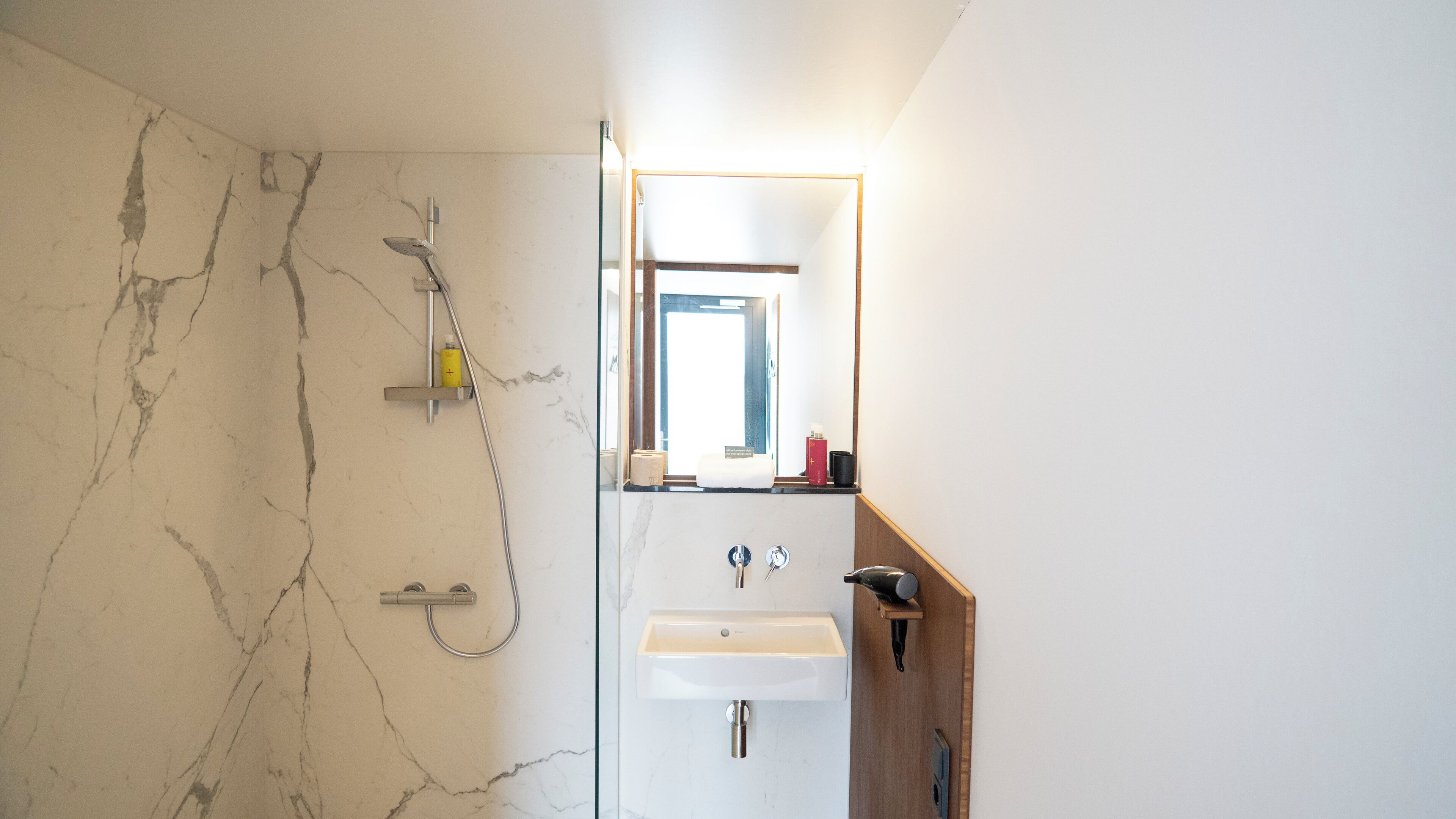 Een badkamer met een marmeren wand, een douche, een wastafel en een spiegel.