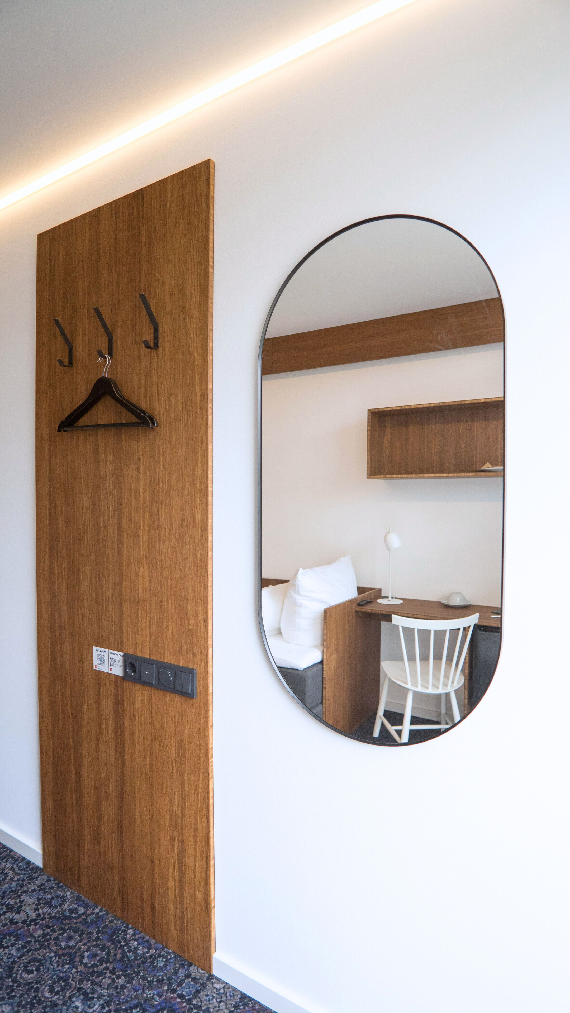 A hotel room with a wooden door, a round mirror, a white chair, a small desk, and a bed with white linens.