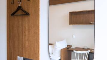 Habitación de hotel con puerta de madera, espejo redondo, silla blanca, escritorio pequeño y cama con ropa blanca.
