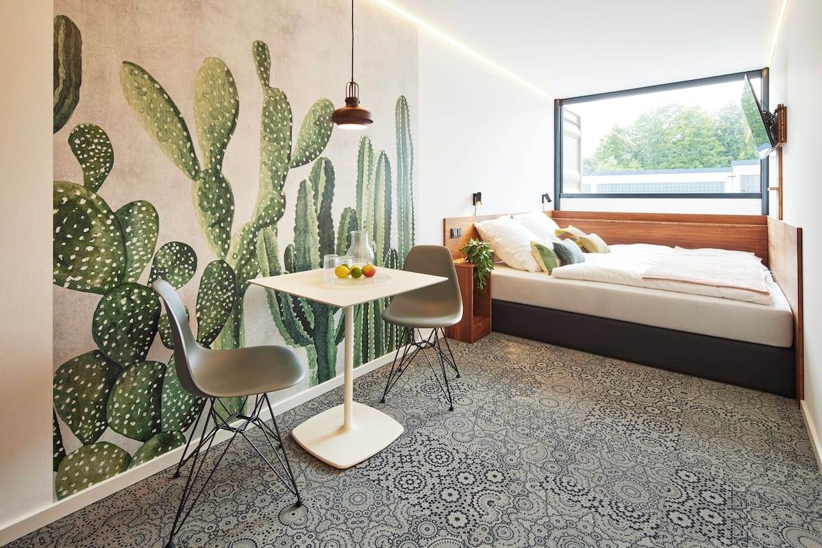 Una habitación moderna con un mural de cactus, una cama con cabecera de madera, una pequeña mesa con sillas y un ventanal que ofrece vistas de la vegetación exterior.