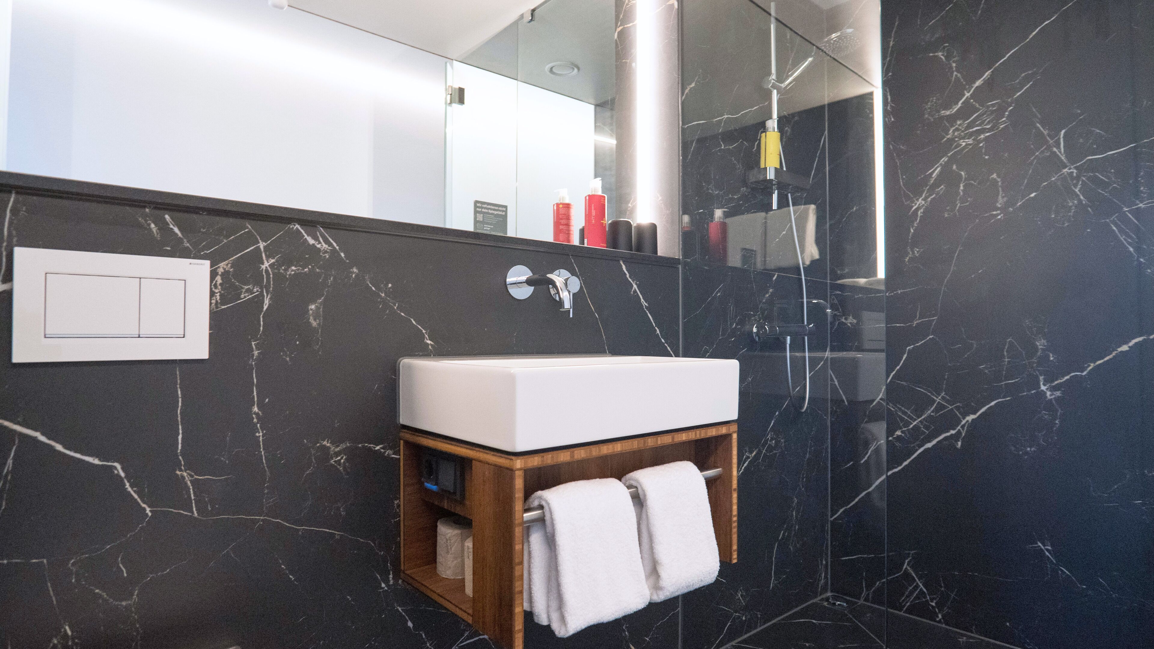 Un baño con lavamanos blanco, paredes de mármol, tocador de madera y un perchero para toallas.