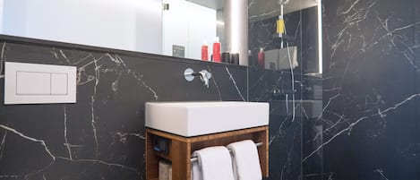 Un baño con lavamanos blanco, paredes de mármol, tocador de madera y un perchero para toallas.