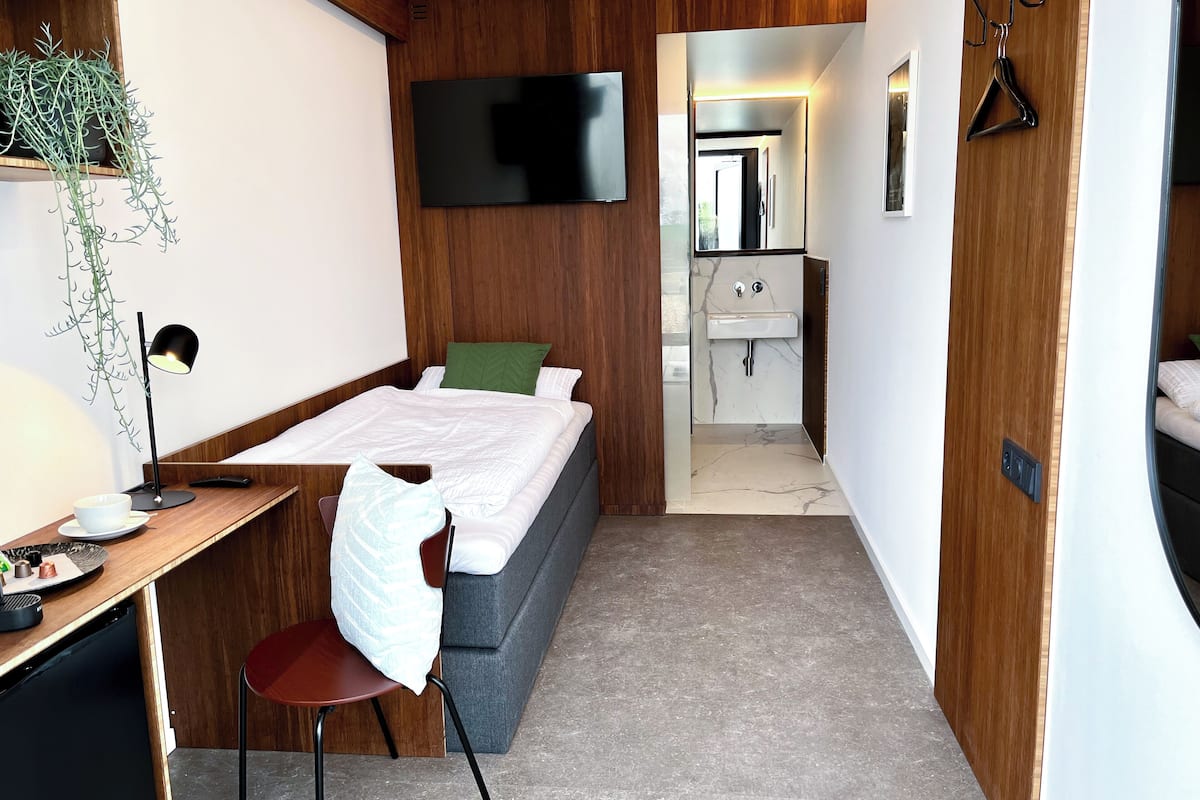 Una habitación de hotel moderna con cama, escritorio, silla, televisión y baño con lavamanos y espejo.