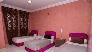 A hotel room with three beds, each with a pink headboard and a white mattress. There is a small wooden nightstand with a phone between the beds. The room has a pink wall and a window with brown curtains. - Hotel Alecanti (Beni Mellal)
