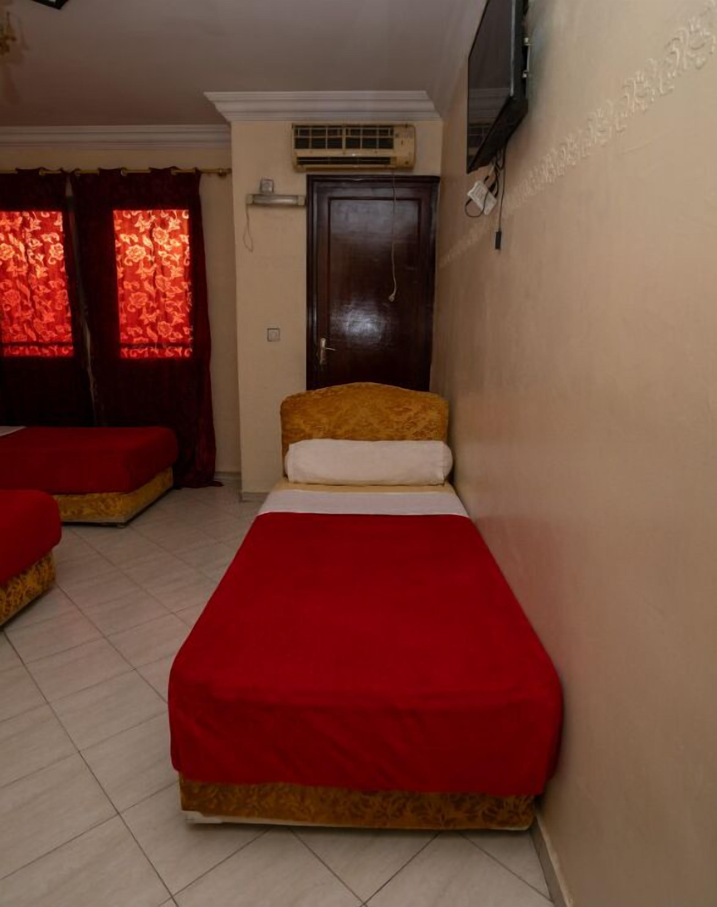 Una habitación pequeña con cama individual, colcha roja, puerta de madera y ventana con cortinas rojas.