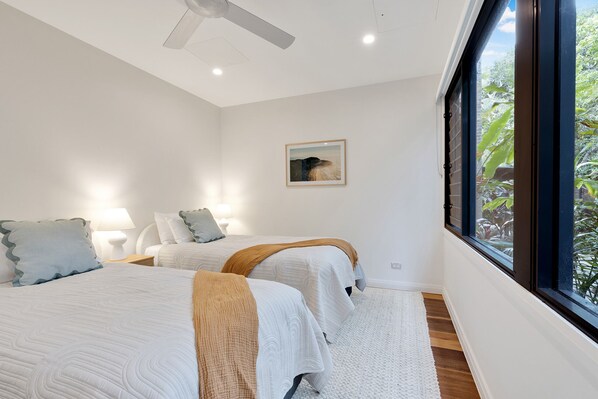 Un dormitorio con dos camas, un ventilador de techo, un cuadro en la pared y una ventana con vistas a la vegetación.