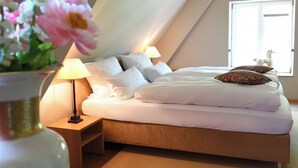 Een slaapkamer met een bed, kussens, een nachtkastje, een lamp en een raam met uitzicht op een wit paard.