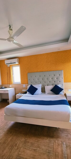 A hotel room with a large bed, a ceiling fan, an air conditioning unit, a desk, and a window. - Limewood Riverside Resort (Ramnagar)