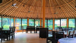 A restaurant with a thatched roof, wooden tables and chairs, and large windows offering a view of greenery. - Limewood Riverside Resort (Ramnagar)