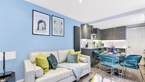 A modern living room with a sofa, a dining area with chairs, and a kitchen in the background. - Tranquility in Bloom: Refreshing 2BR Apartment (London)
