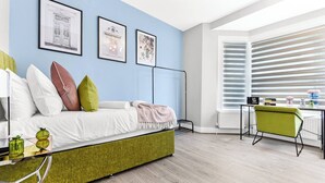 A modern bedroom with a green sofa bed, a desk with a chair, and a large window with blinds. - Tranquility in Bloom: Refreshing 2BR Apartment (London)