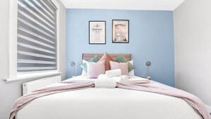 A neatly made bed with a white and pink bedspread, multiple pillows, and a headboard. There are two framed pictures on the wall above the bed. - Tranquility in Bloom: Refreshing 2BR Apartment (London)