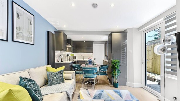 A modern living room with a sofa, a coffee table, and a dining area with chairs and a table. There is a kitchen in the background, a large window with blinds, and a potted plant. - Tranquility in Bloom: Refreshing 2BR Apartment (London)