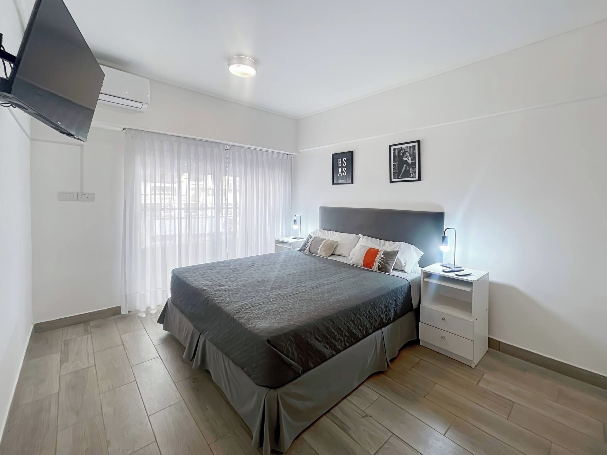 A modern bedroom with a large bed, bedside tables, a flat-screen TV, and a window with sheer curtains.