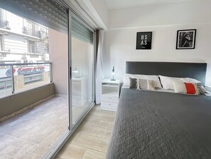 A modern bedroom with a large bed, a nightstand, a bedside table, and a view of a cityscape through a sliding glass door. - Modern Studio With Amenities in the Heart of the City (Buenos Aires)