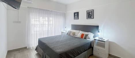 A modern bedroom with a large bed, bedside tables, a flat-screen TV, and a window with sheer curtains.