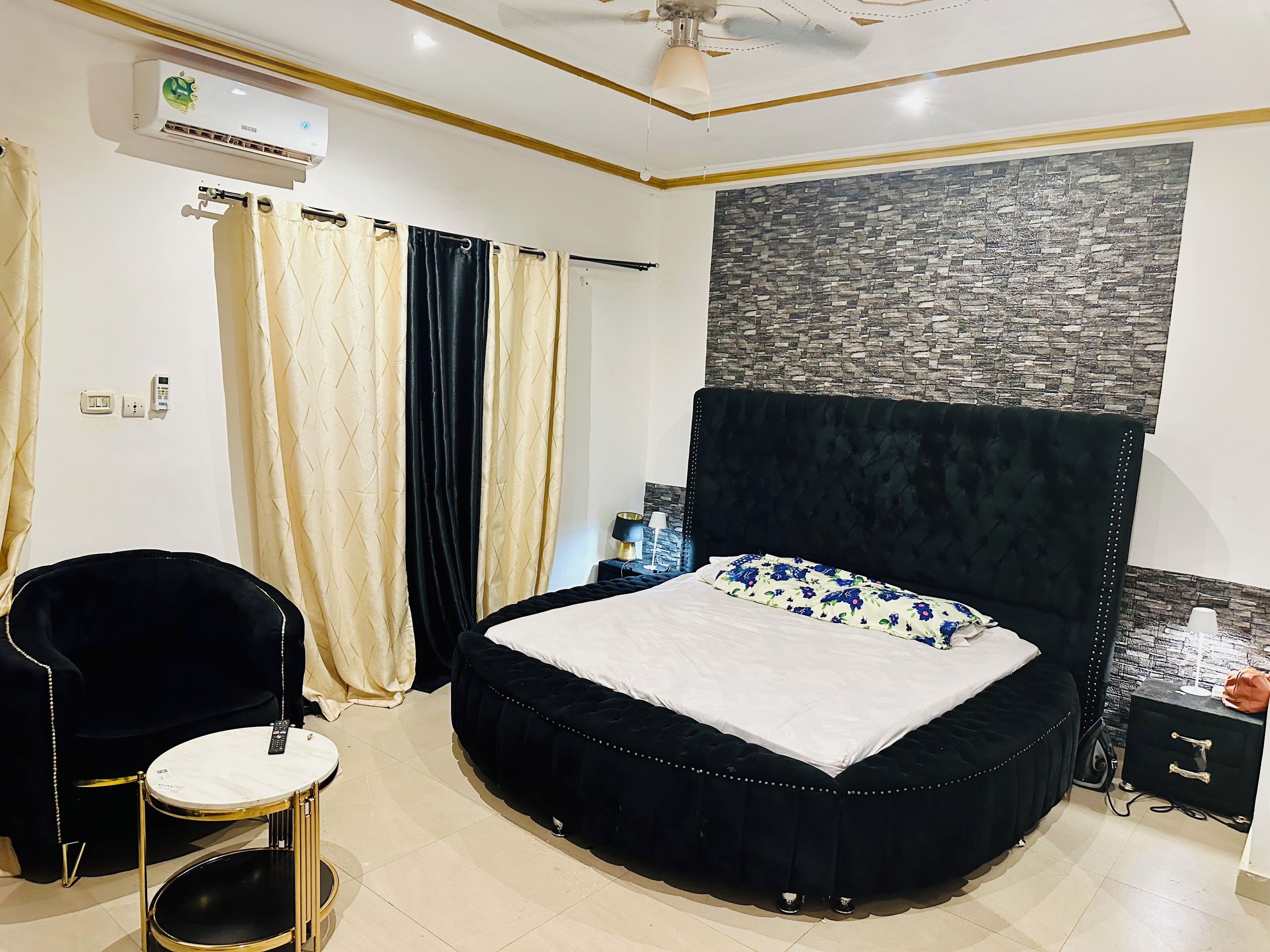 A bedroom with a round bed, a black upholstered chair, a small round table, and a wall-mounted air conditioner.