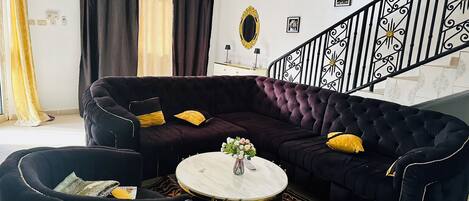 Una sala de estar con un sofá oscuro, una mesa redonda de centro y una escalera con barandillas decorativas.