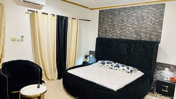 Une chambre à coucher avec un lit rond, un fauteuil noir recouvert de tissu, une petite table ronde et un climatiseur fixé au mur.