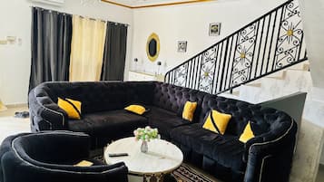 Un salon avec un canapé d’angle noir, une table basse ronde et un escalier doté de rampes noires décoratives.