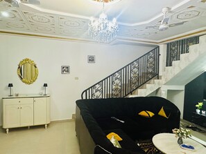 A living room with a staircase, a chandelier, a white cabinet, and a black sofa. - Villa Amina Splendid Elegant Structure (Ouagadougou)