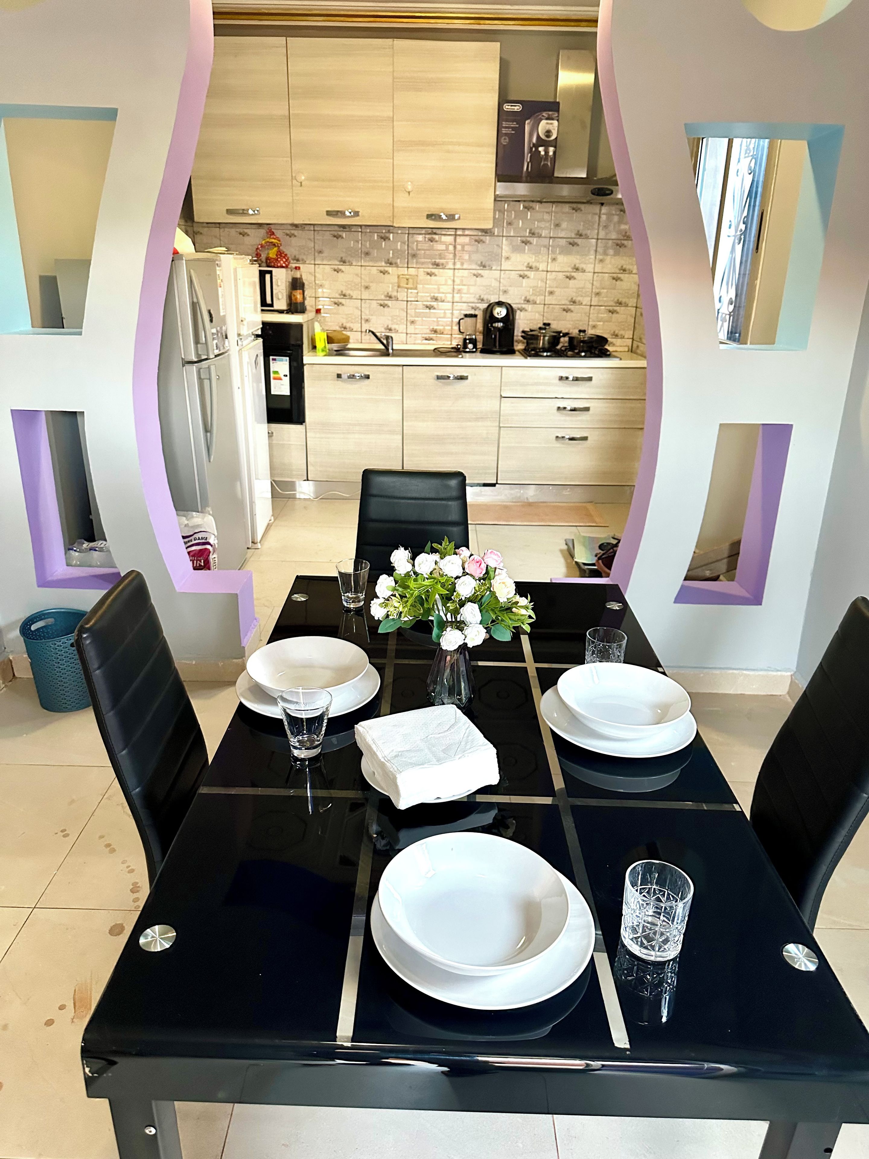 Un coin repas avec une table noire dressée pour quatre personnes, des assiettes blanches, des verres et un vase rempli de fleurs.