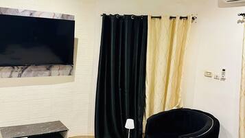 Une chambre moderne équipée d’un téléviseur à écran plat, d’un fauteuil noir, d’une petite table ronde avec une lampe, d’un tapis noir et doré à motifs et d’un ventilateur de plafond.