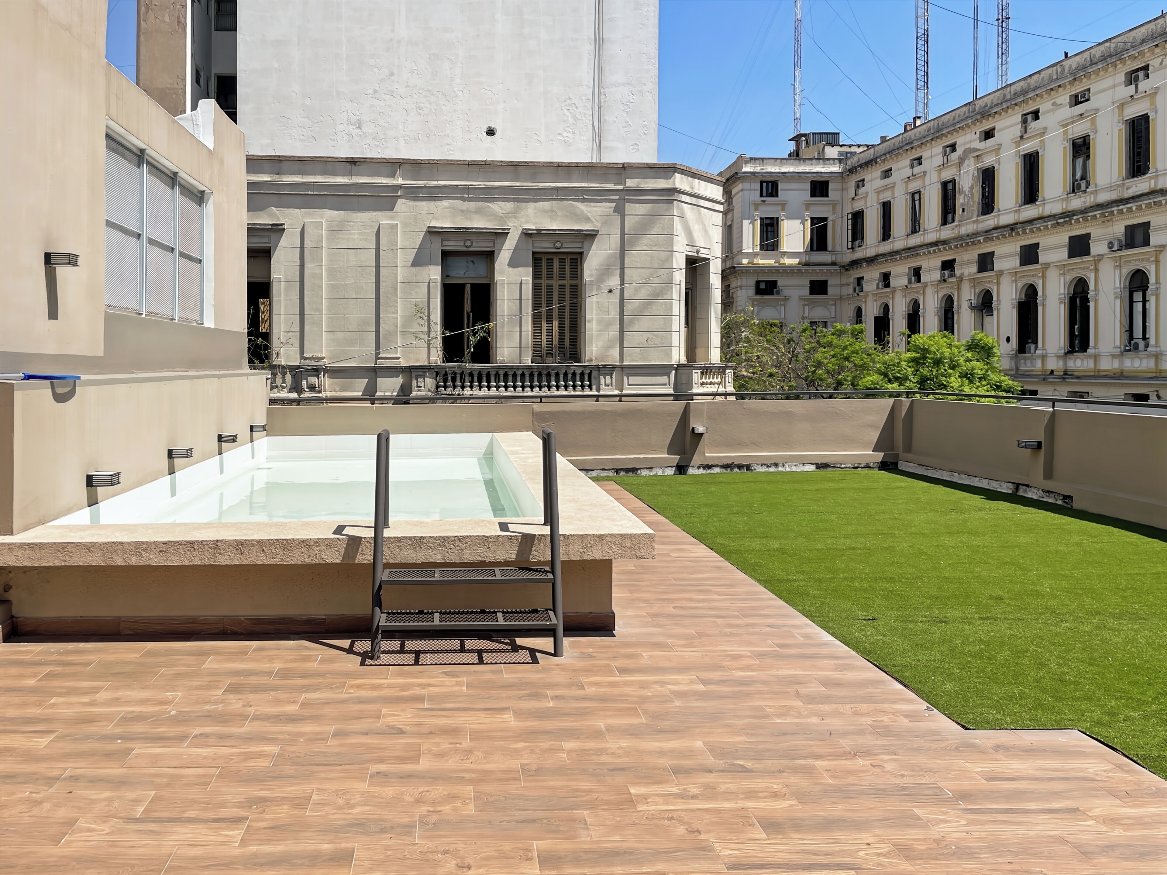A rooftop pool with a wooden deck, surrounded by a well-maintained lawn and classical architecture.