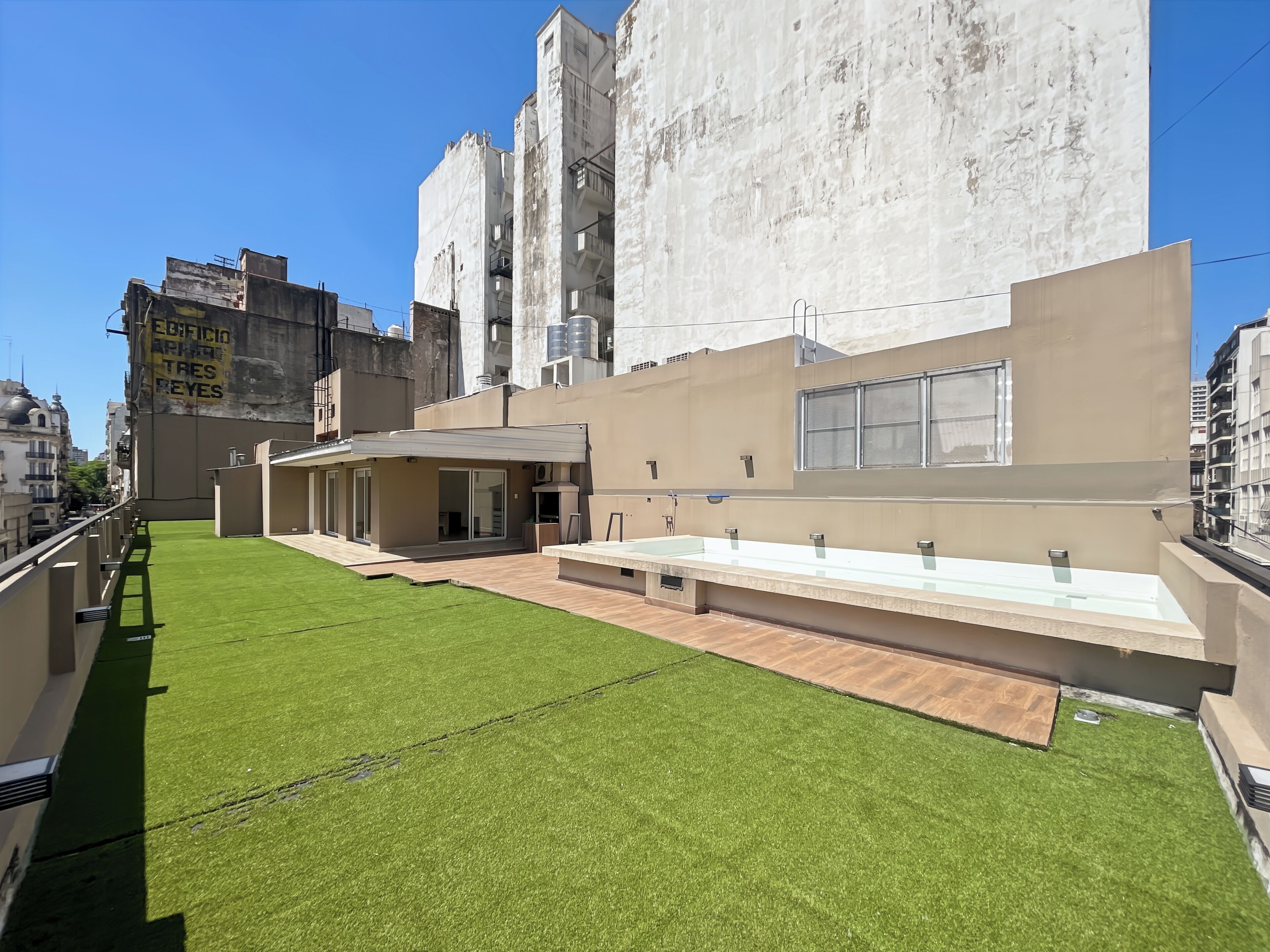 A modern building with a flat roof, a large outdoor area with artificial grass, and a long, narrow pool.