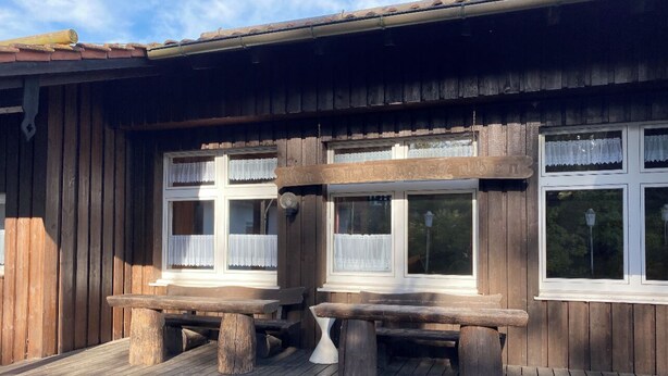 Un bâtiment en bois doté de deux fenêtres, chacune ornée d’un cadre blanc et de rideaux en dentelle. Des bancs en bois sont placés devant les fenêtres.