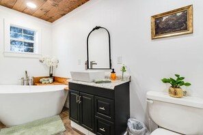 A modern bathroom with a freestanding bathtub, a sink with a mirror above it, and a toilet. There is a window with a view of trees, and a framed picture on the wall.