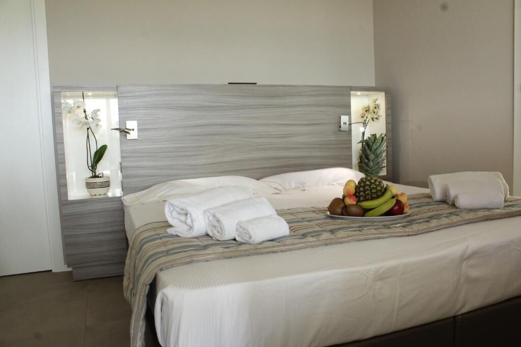 Cama arrumada com roupa de cama branca, uma cesta de frutas sobre a cama e uma janela com uma planta de orquídea.
