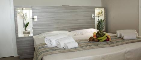 Cama arrumada com roupa de cama branca, uma cesta de frutas sobre a cama e uma janela com uma planta de orquídea.