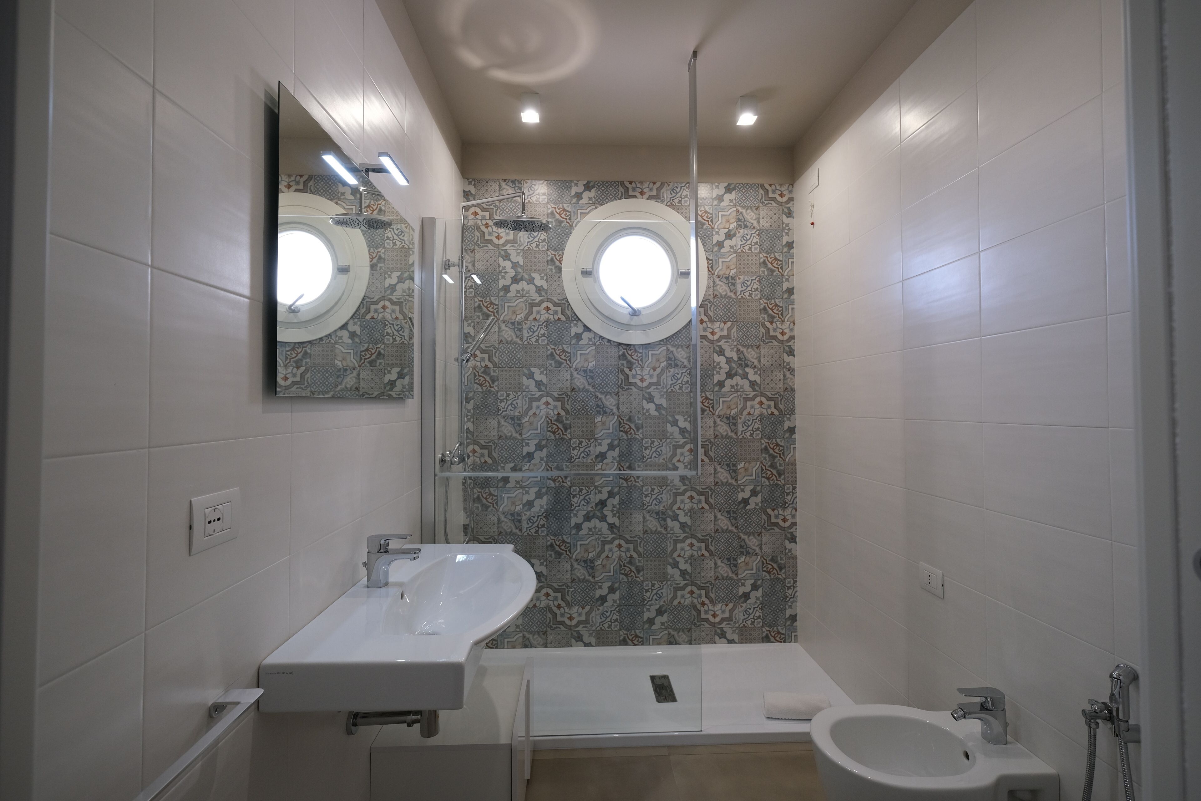 A modern bathroom with a large mirror, two sinks, a bidet, and a shower area.