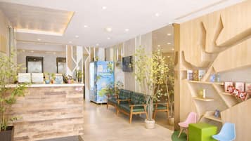 Un área de recepción con un mostrador de madera, sillas verdes y rosas, un sofá verde y un televisor montado en la pared.
