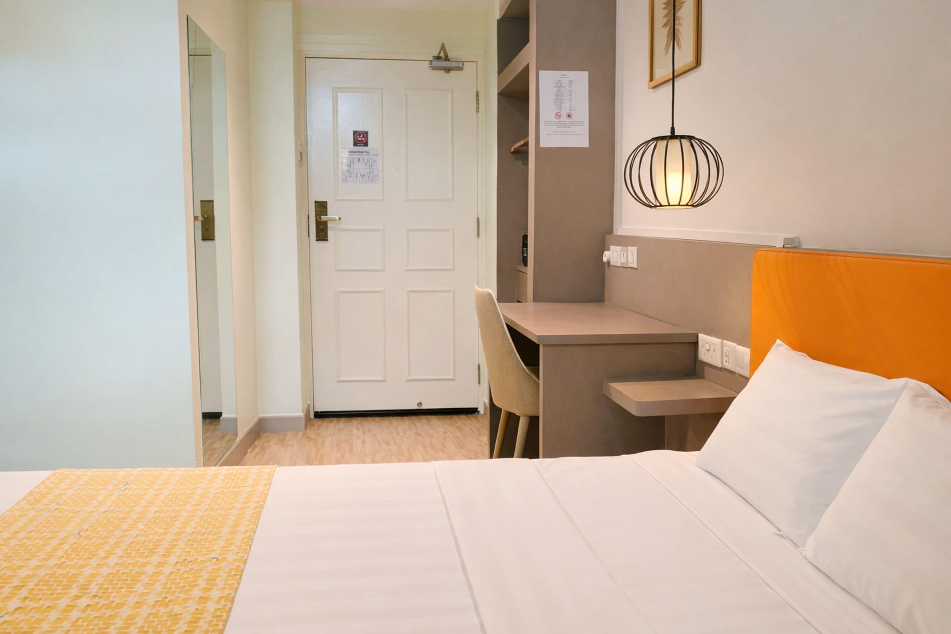 A hotel room with a bed, a desk, and a chair. There is a door with a sign on it, and a wall-mounted light fixture.