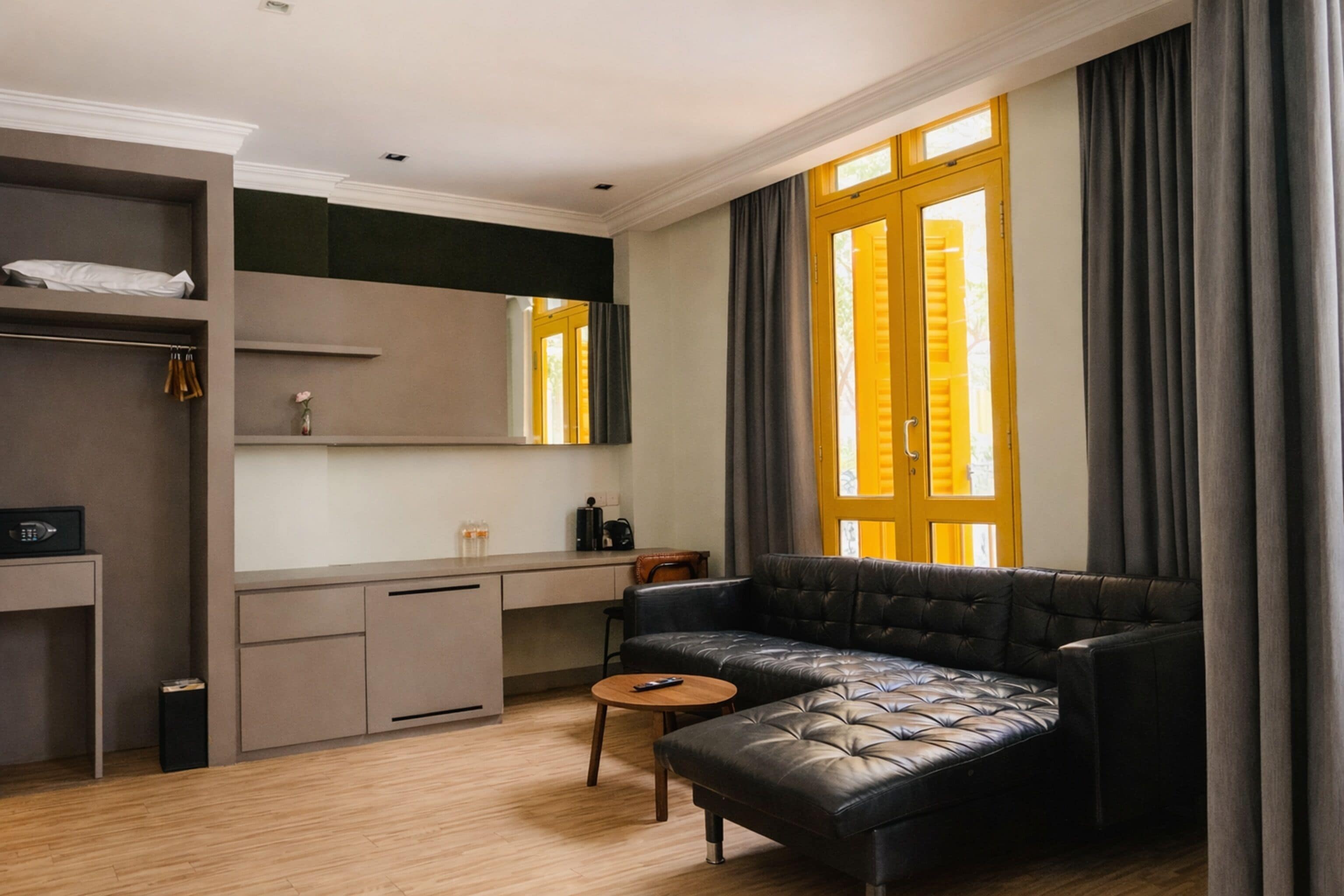 A modern living room with a leather sofa, a small round table, a desk with a chair, a wardrobe, and a large window with curtains.