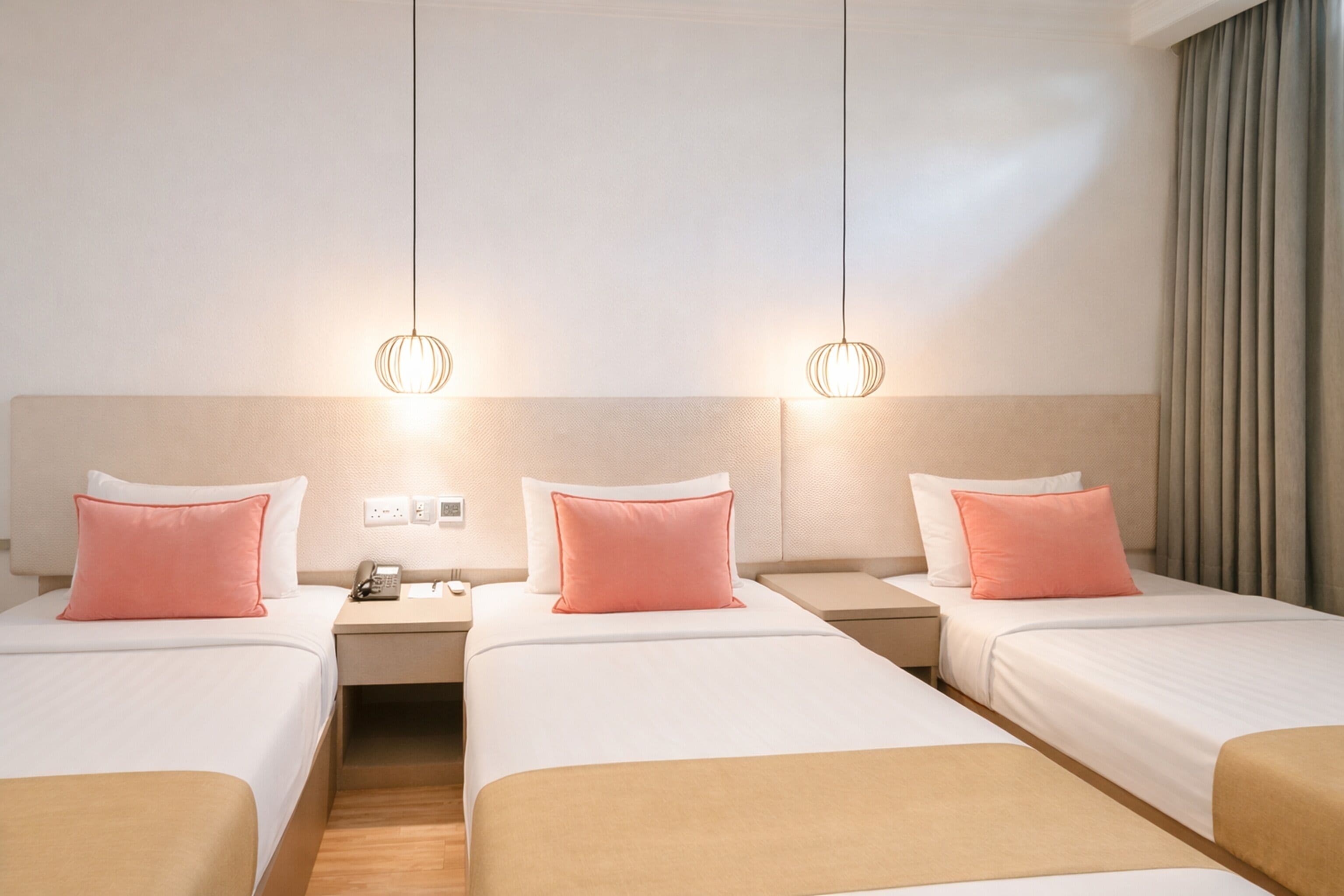 A hotel room with three beds, each with a white and pink pillow, and a beige headboard. There are two bedside tables with a phone and a lamp on each. The room has a wooden floor and a curtain on the window.