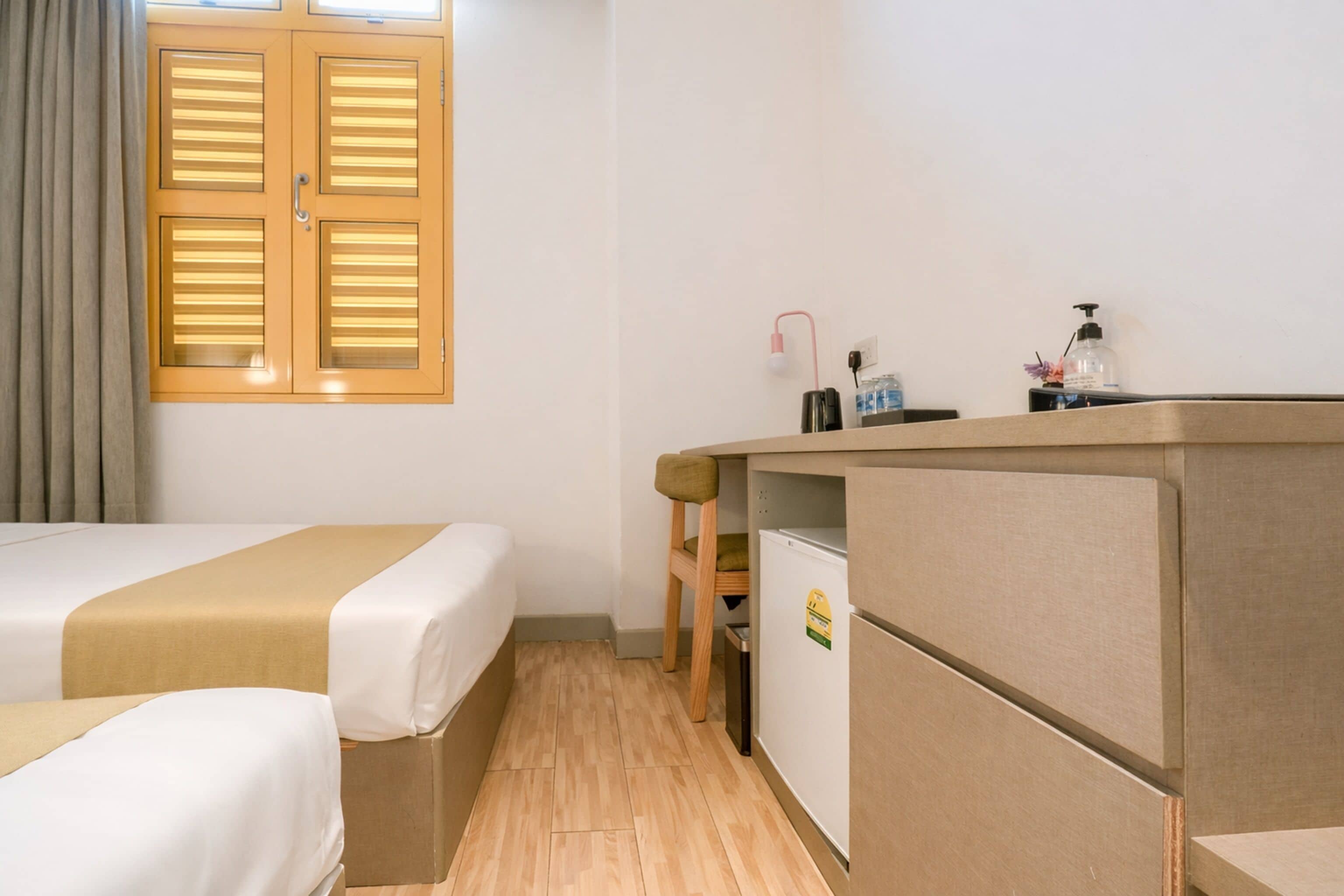 Quarto de hotel com duas camas, uma pequena cozinha compacta, uma cadeira de madeira e uma janela com persianas.