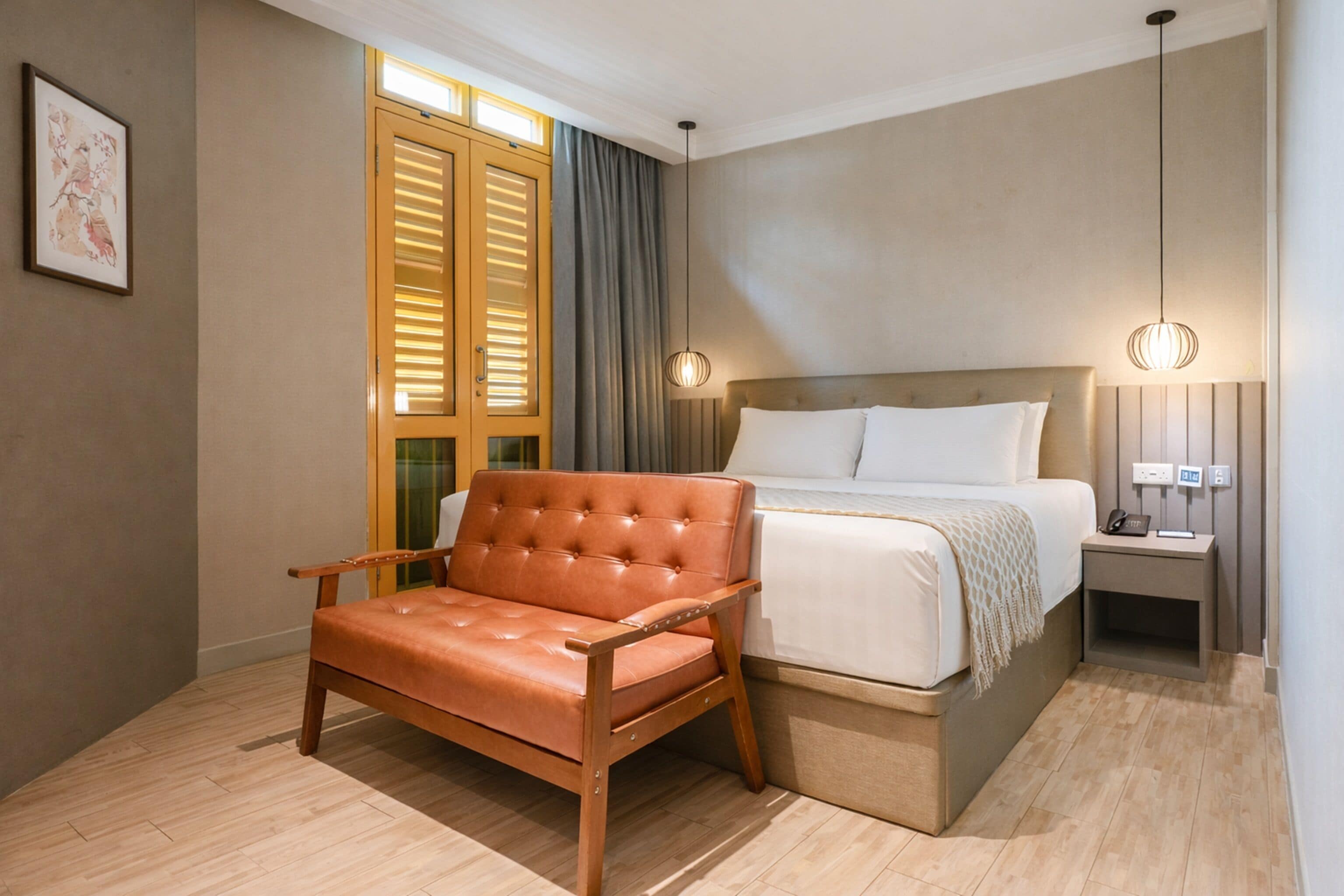 A hotel room with a bed, a leather chair, a bedside table with a lamp, and a window with blinds.