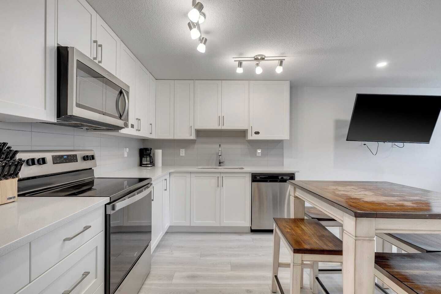 Une cuisine moderne avec des meubles blancs, des appareils électroménagers en acier inoxydable, une table à manger en bois et un téléviseur à écran plat fixé au mur.