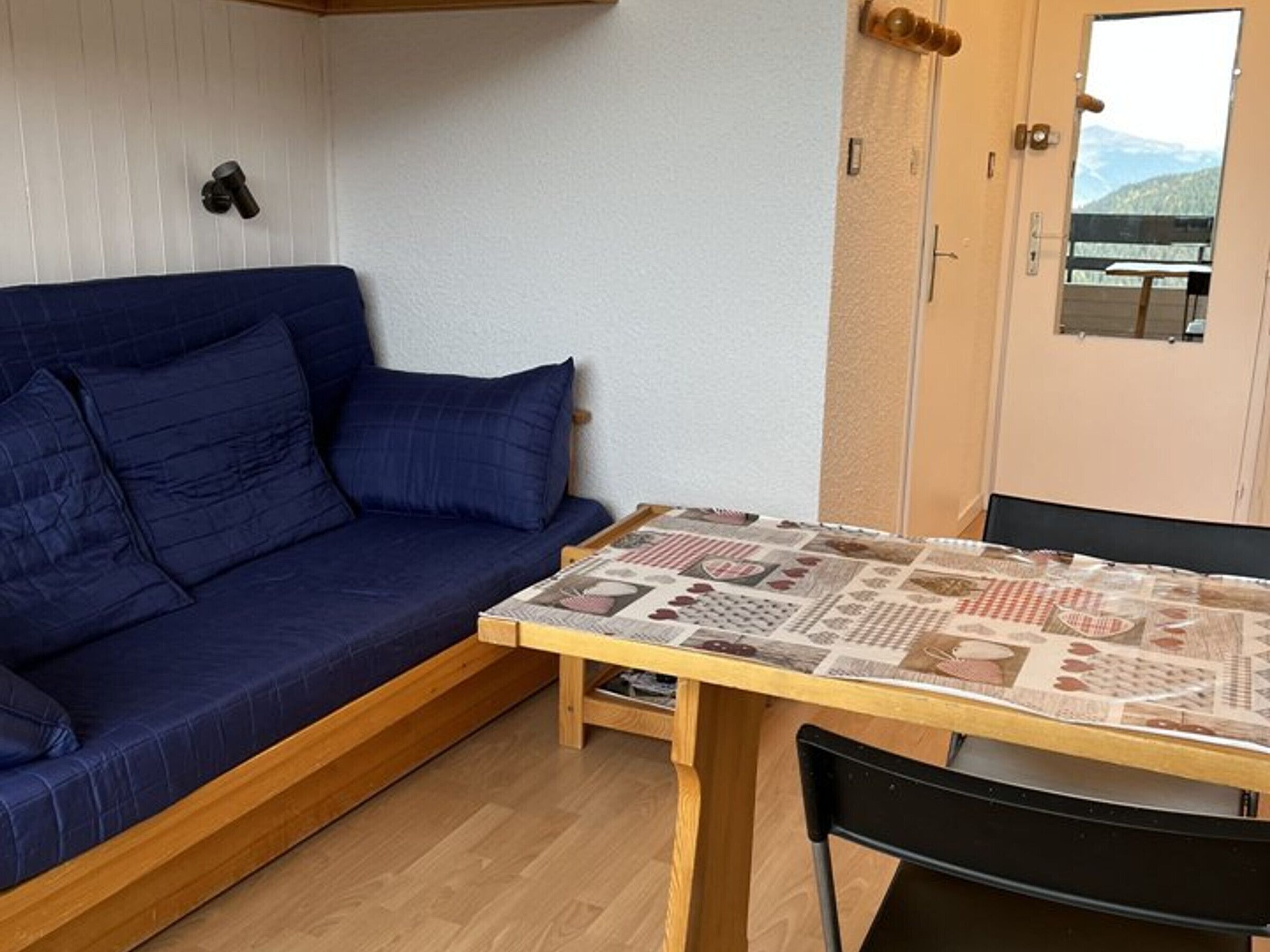 Un espace de vie compact avec un canapé bleu, une table en bois recouverte d’une nappe à motifs et une porte donnant sur une vue sur les montagnes.