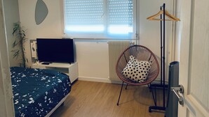 Makuuhuoneessa on sänky, tuoli, televisio, patteri ja ikkuna, jossa on sälekaihtimet.