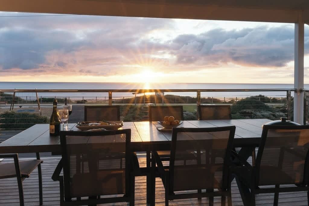 A wooden deck with a table and chairs, overlooking a beach at sunset.