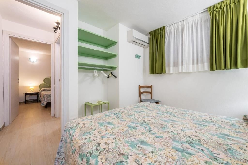 A bedroom with a floral bedspread, a green shelf unit, a chair, and a window with green and white curtains.