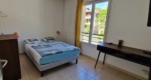 Quarto com cama, escrivaninha, radiador e janela com vista para edifícios.