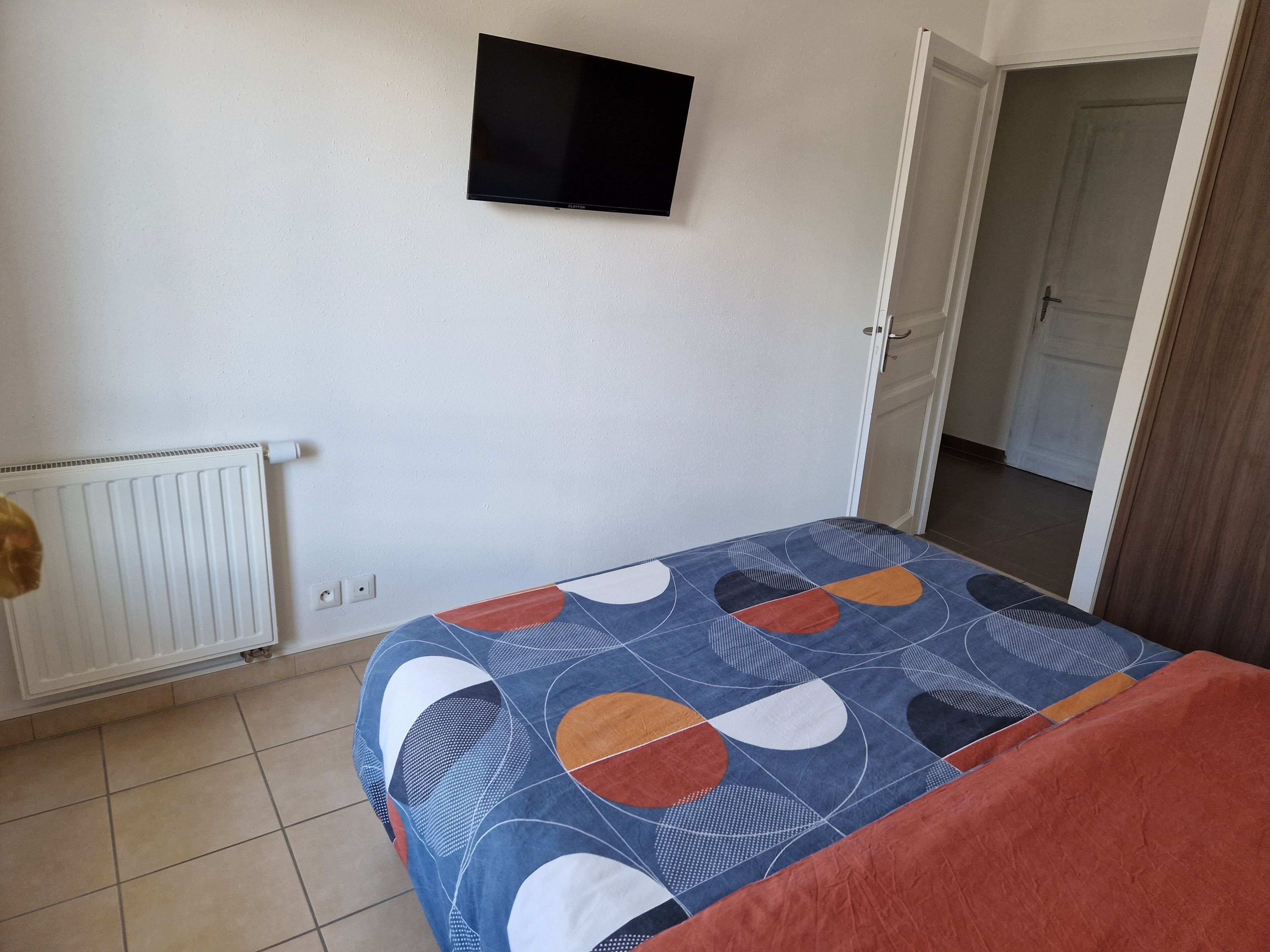 Un dormitorio con cama, un radiador, un televisor en la pared y una puerta que da a otra habitación.