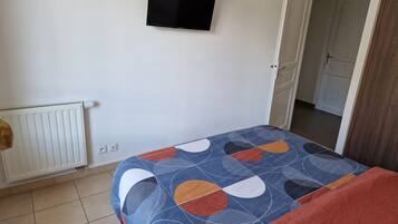 Un dormitorio con cama, un radiador, un televisor en la pared y una puerta que da a otra habitación.
