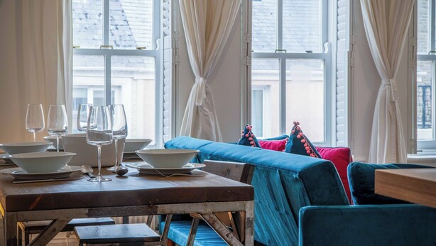 Ein Essbereich mit einem Holztisch, gedeckt für eine Mahlzeit, einem blauen Sofa mit bunten Kissen und großen Fenstern mit Vorhängen.